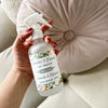 Hand holding a bottle of Dot & Lil air freshener with a floral label against a neutral background.